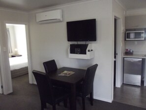 A compact hotel room with a dining table, chairs, a flat-screen TV mounted on the wall, a microwave, and a compact kitchenette.