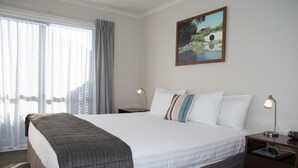 Ein Hotelzimmer mit einem großen Bett, einem Nachttisch mit Lampe, einem Fenster mit Vorhängen und einem Bild an der Wand.