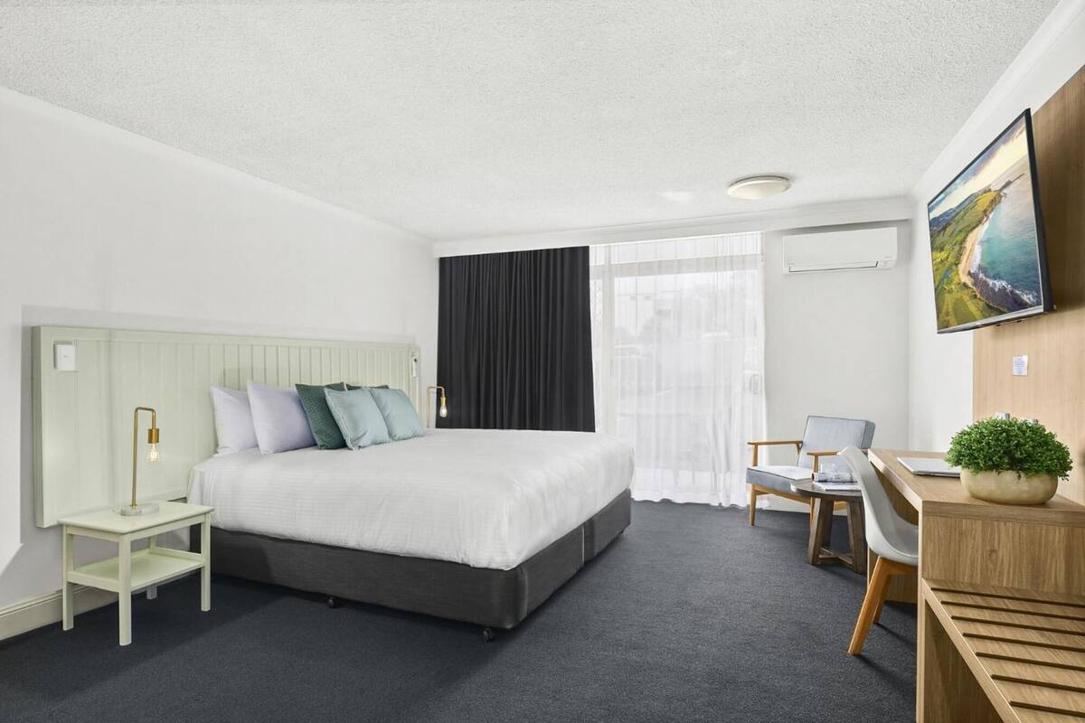 Une chambre d’hôtel avec un lit, un bureau, une chaise, une petite table, une plante et un tableau au mur.