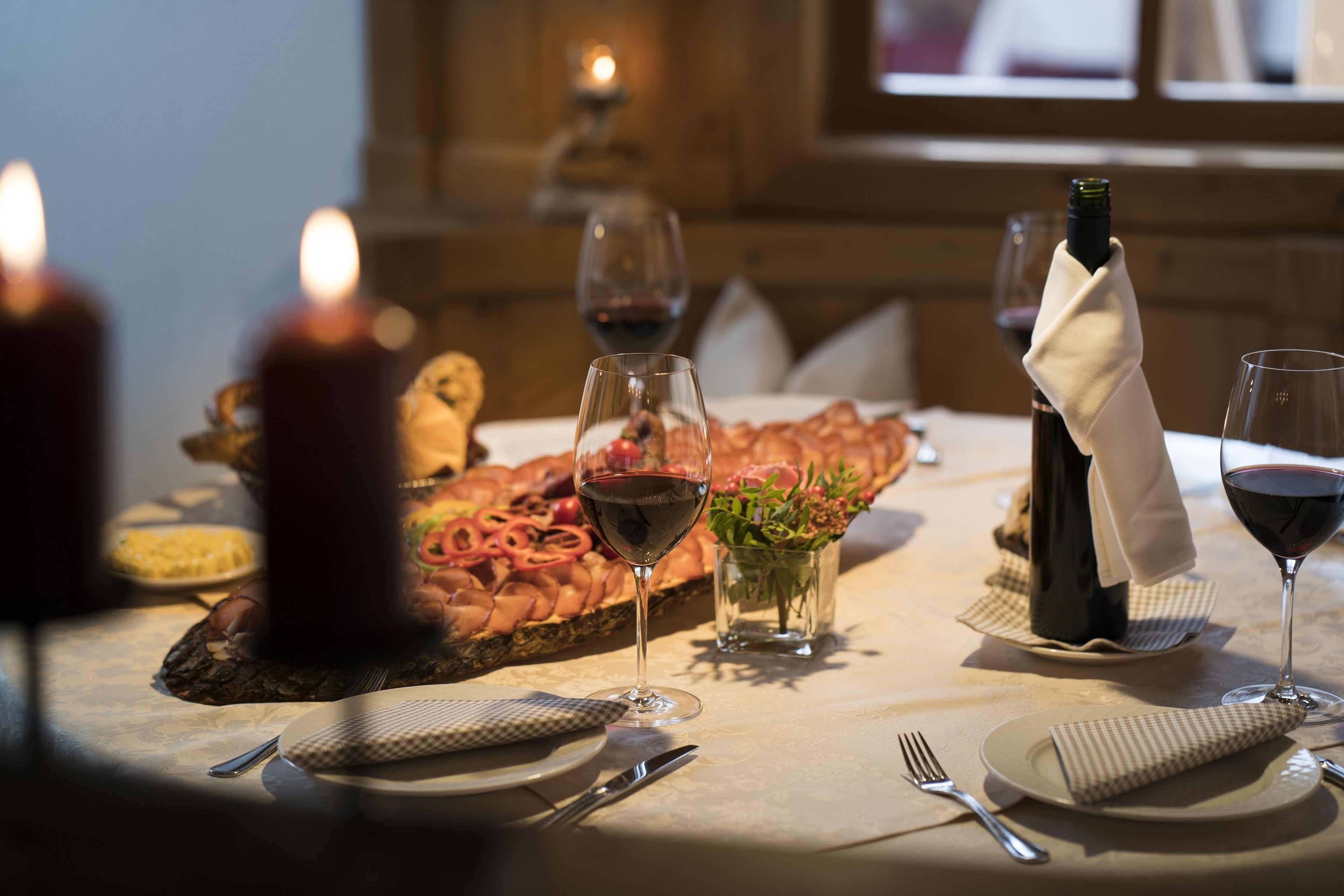A dining table set with plates, cutlery, glasses, and a bottle of wine, with a candle and a vase of flowers in the background.