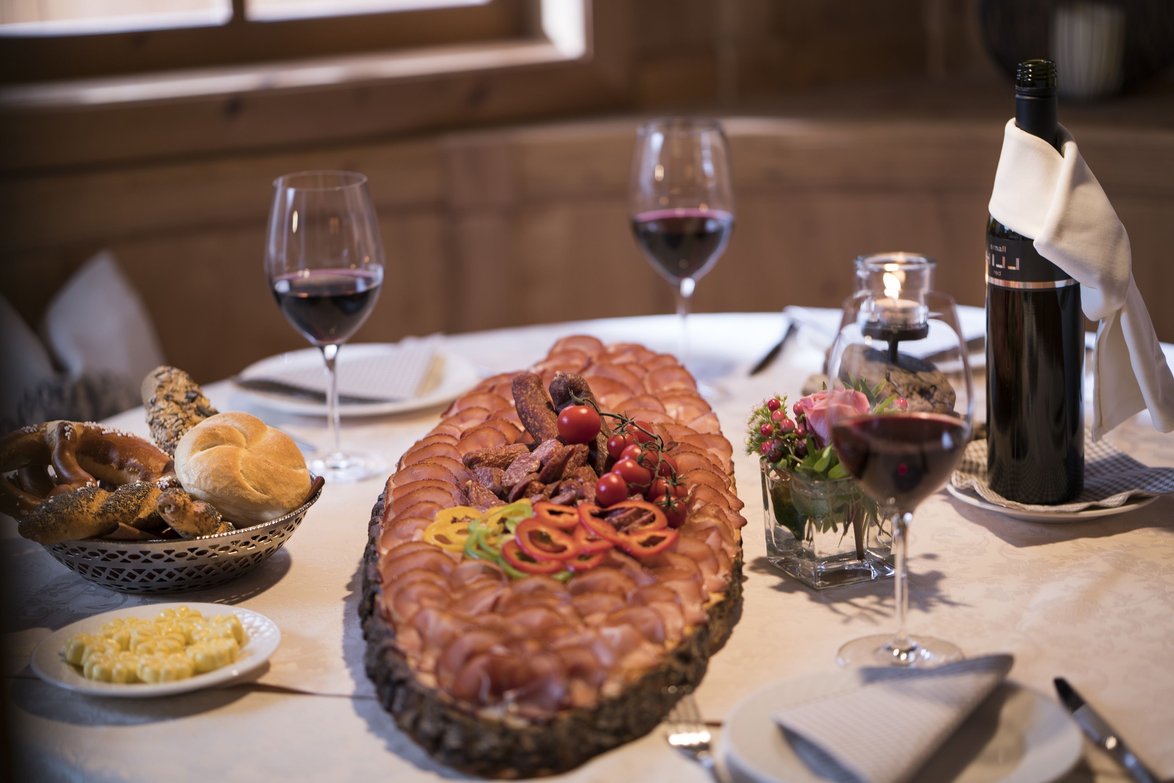 A dining table with a large meat dish, two glasses of red wine, a bottle of wine, a bowl of pretzels, a plate of macaroni, and a small vase with flowers.