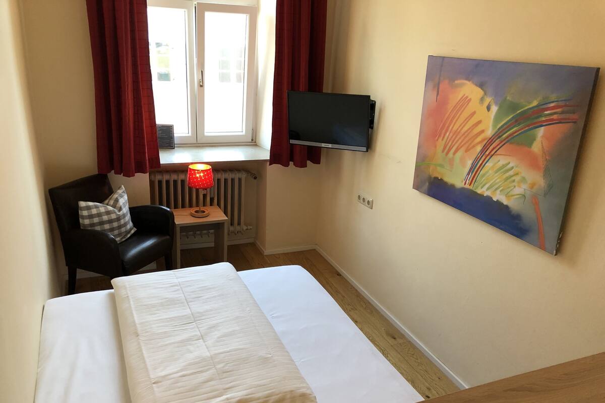 Ein kleines Hotelzimmer mit einem Bett, einem Schreibtisch mit Stuhl, einem Heizkörper, einem an der Wand befestigten Fernseher und einem Fenster mit roten Vorhängen.