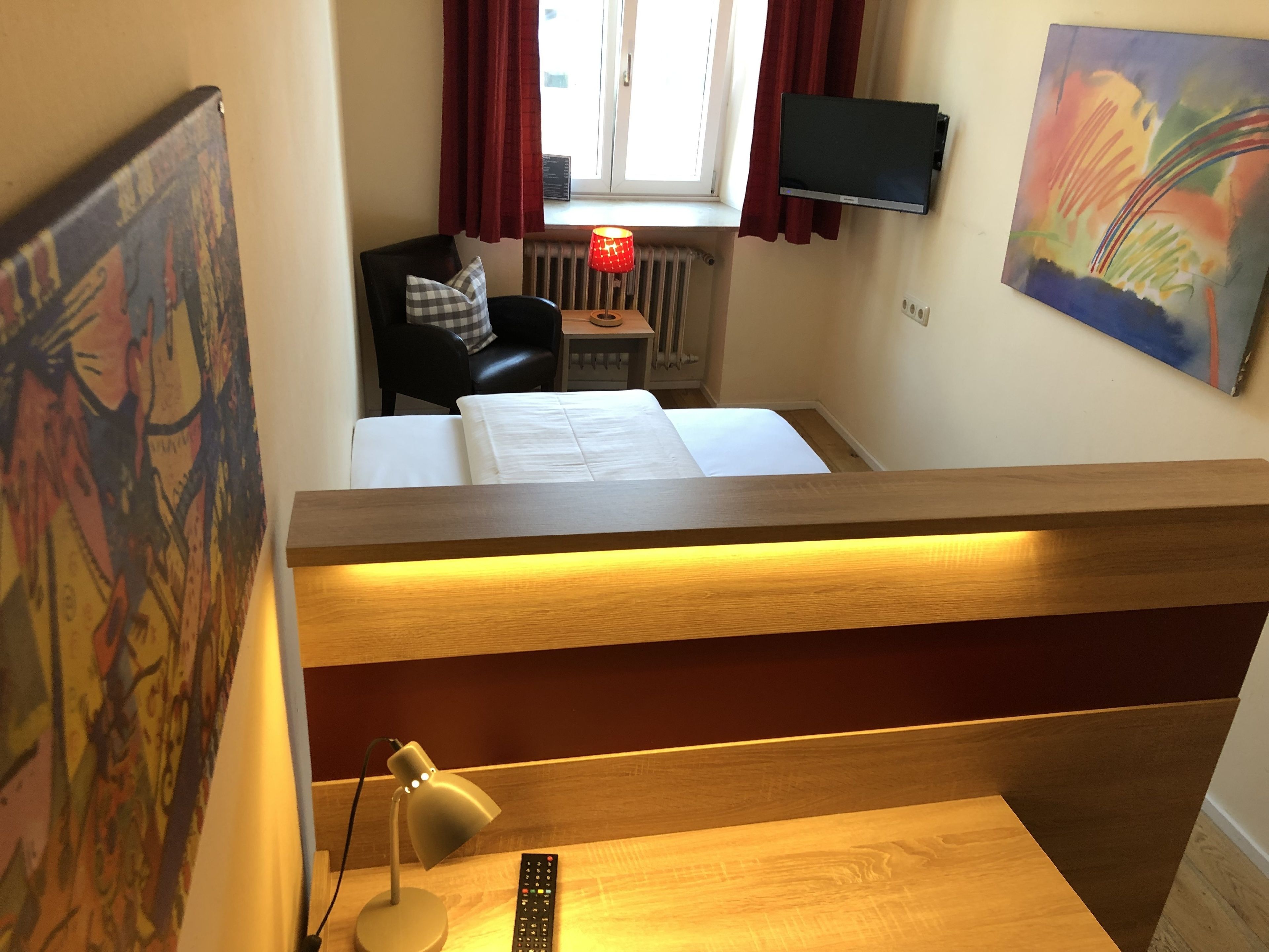 A hotel room with a bed, a desk with a lamp, a chair, a TV, and a painting on the wall.