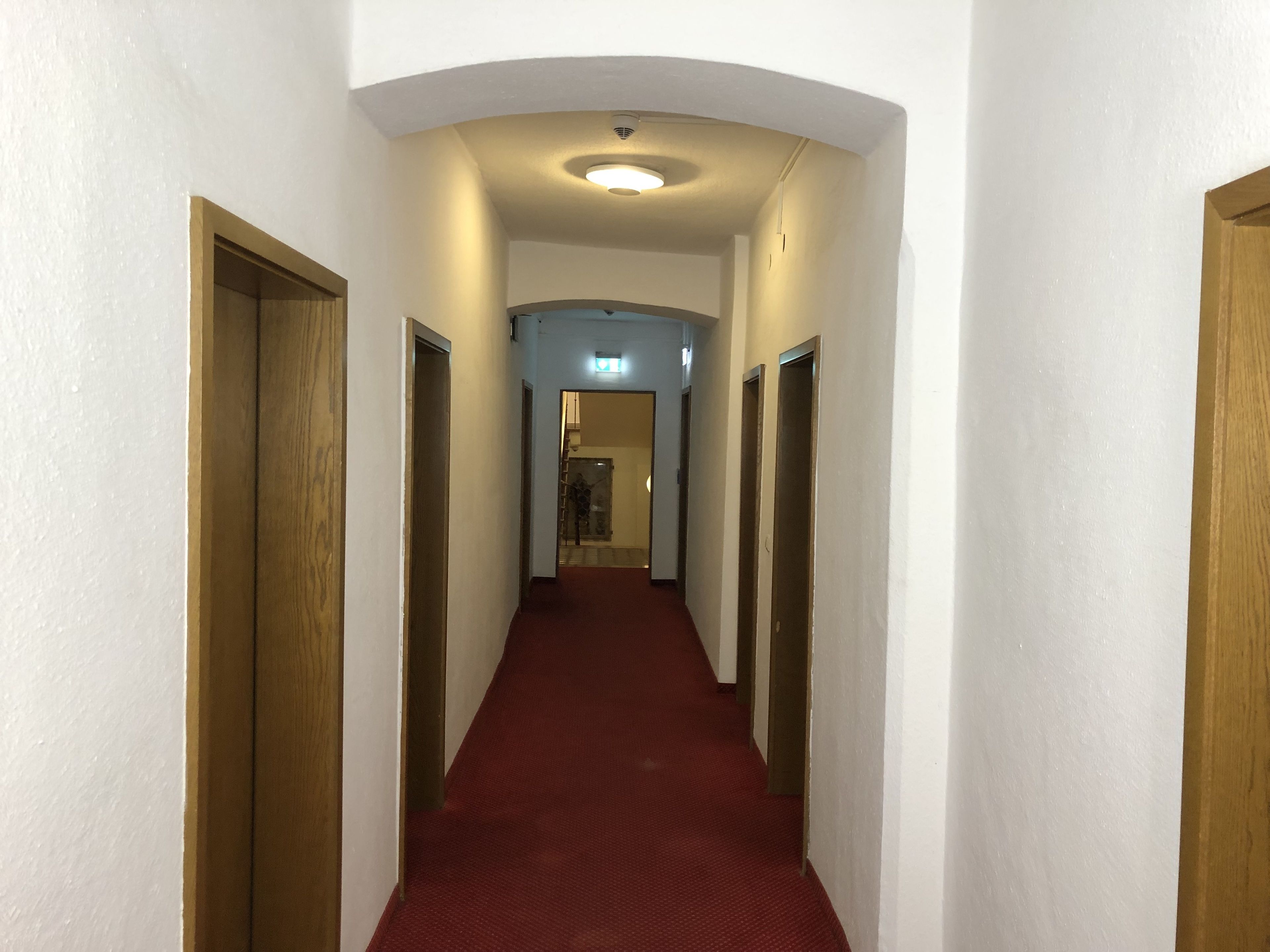 Un pasillo estrecho con alfombra roja, paredes blancas y puertas de madera a ambos lados.