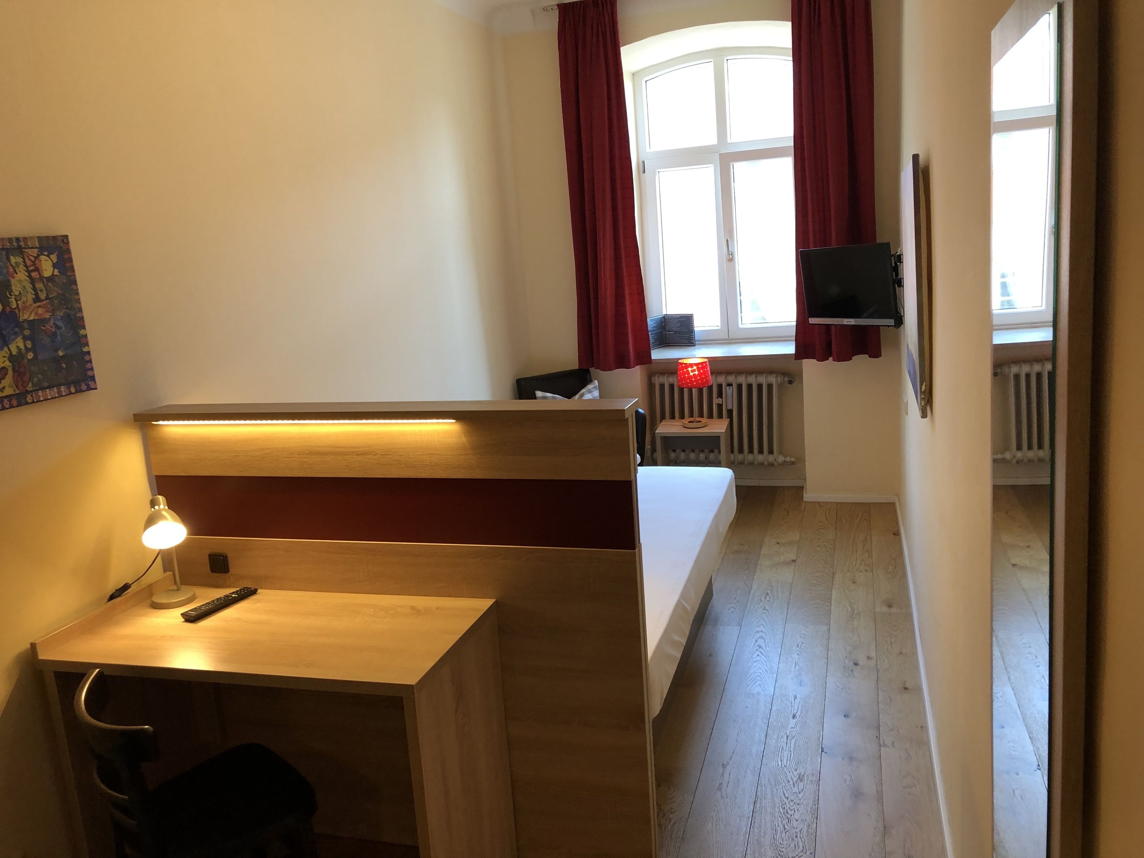 Ein Zimmer mit einem Holztisch, einem Stuhl, einem Bett, einem Fernseher und einem fenster mit Vorhängen.