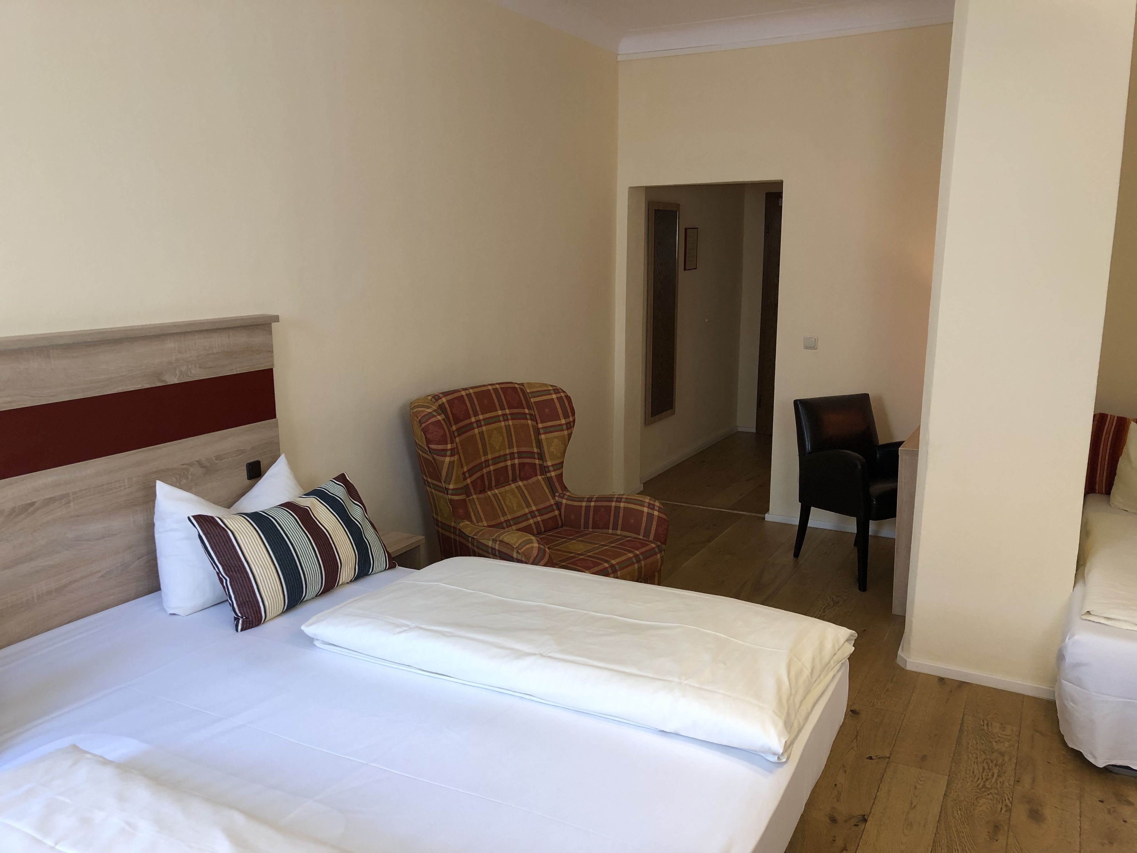 A hotel room with a bed, a plaid armchair, a black chair, and a wooden floor.