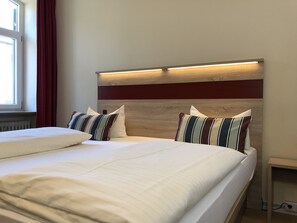 A neatly made bed with white linens and striped pillows, a wooden headboard with integrated lighting, and a window with red curtains. - Hotel Gasthof Flötzinger Bräu (Rosenheim)