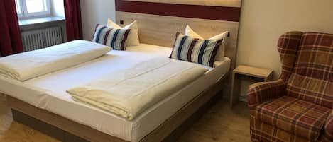 Una cama doble con dos almohadas, cabecera con iluminación integrada, mesita de noche de madera y un sillón a cuadros.