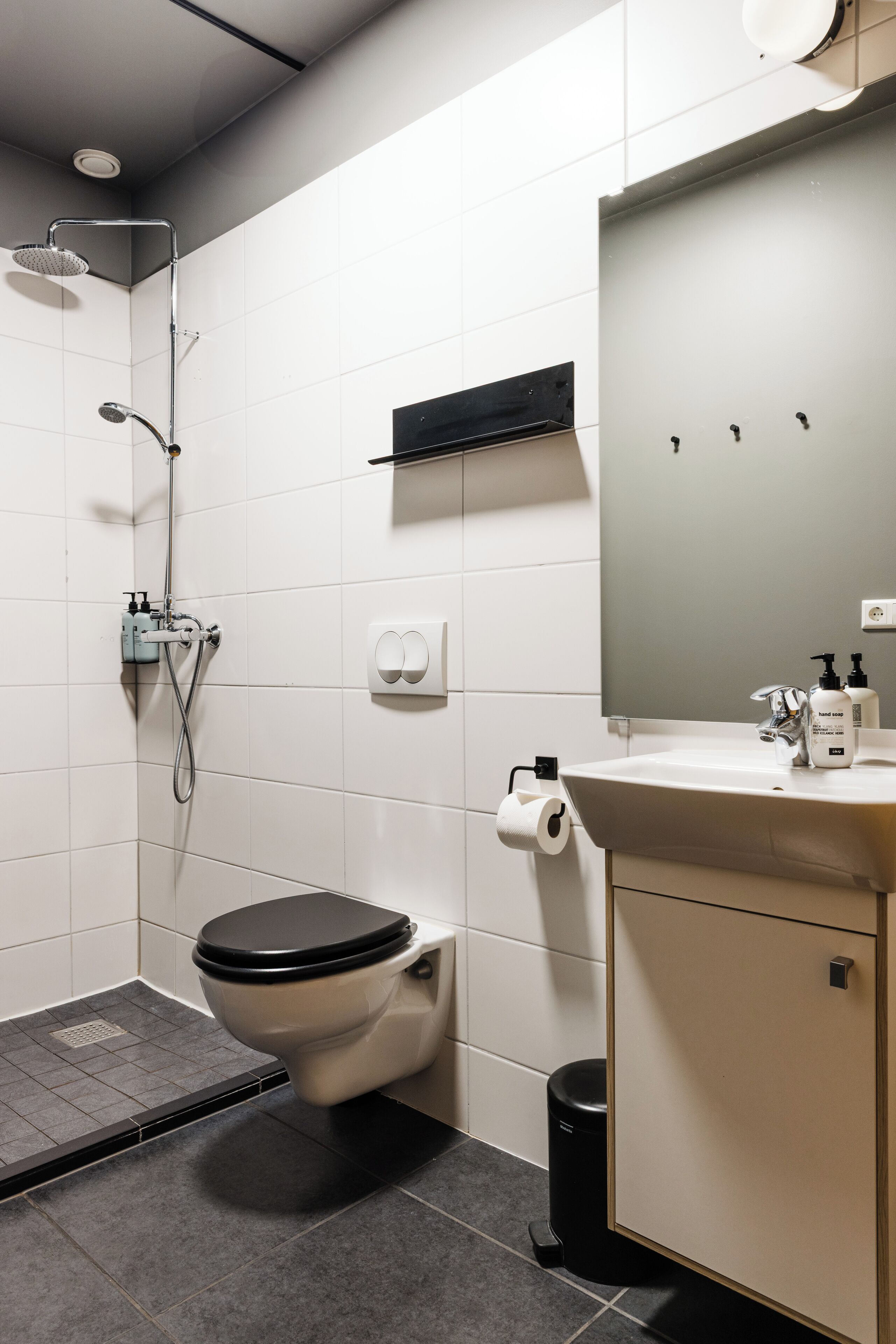 Ein modernes Badezimmer mit Toilette, Waschbecken, Spiegel, Dusche und einem Mülleimer.