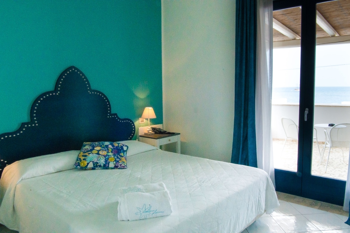 A bed with a headboard, a pillow, a bedside table with a lamp, and a view of the beach through an open door.