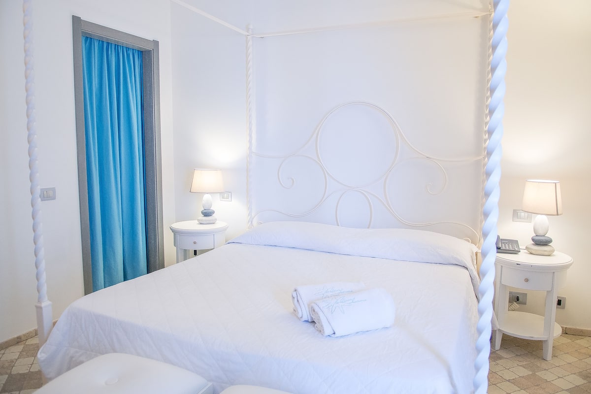 A four-poster bed with white bedding, two bedside tables with lamps, and a blue curtain.