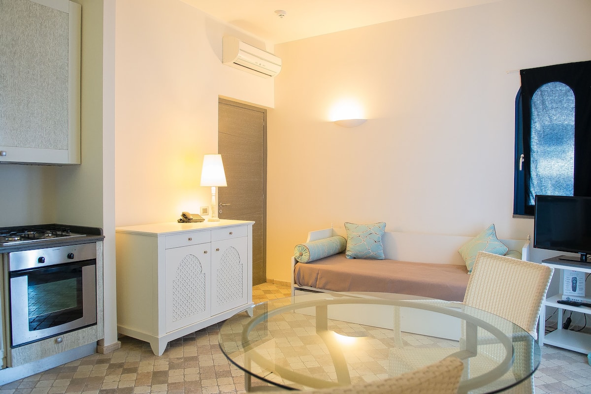 A compact living space with a kitchenette, a bed with pillows, a glass table, and a television.