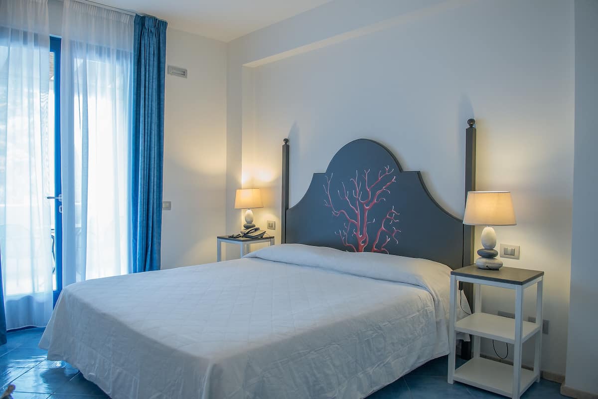 A neatly made bed with white bedding, two bedside tables with lamps, a wall-mounted decorative headboard, and a window with blue curtains.