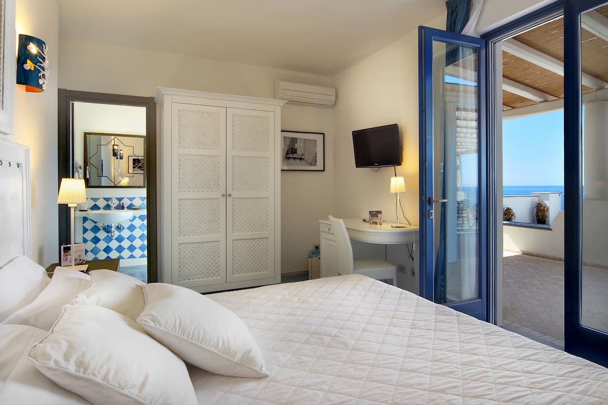 A hotel room with a bed, a desk, a TV, a wardrobe, and a balcony with a view of the sea.