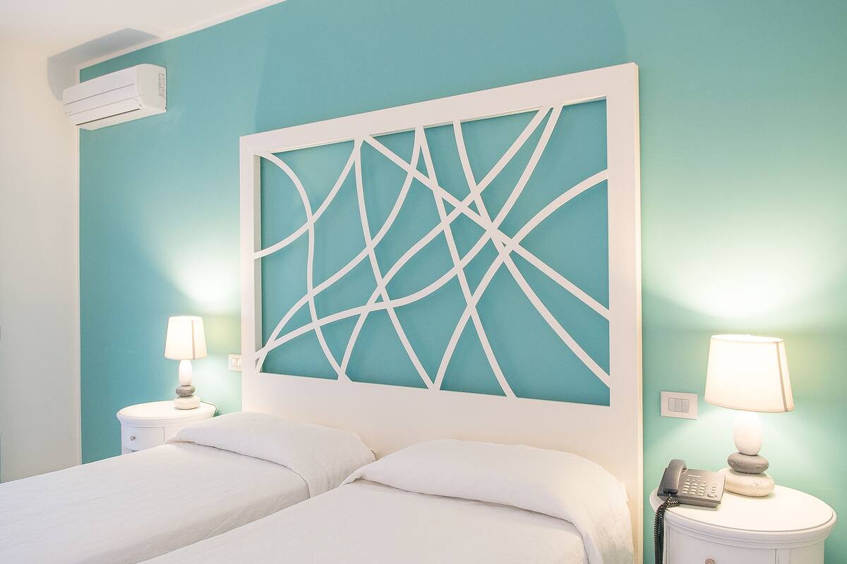 A hotel room with a bed, two bedside tables, a wall-mounted air conditioner, and a decorative headboard.