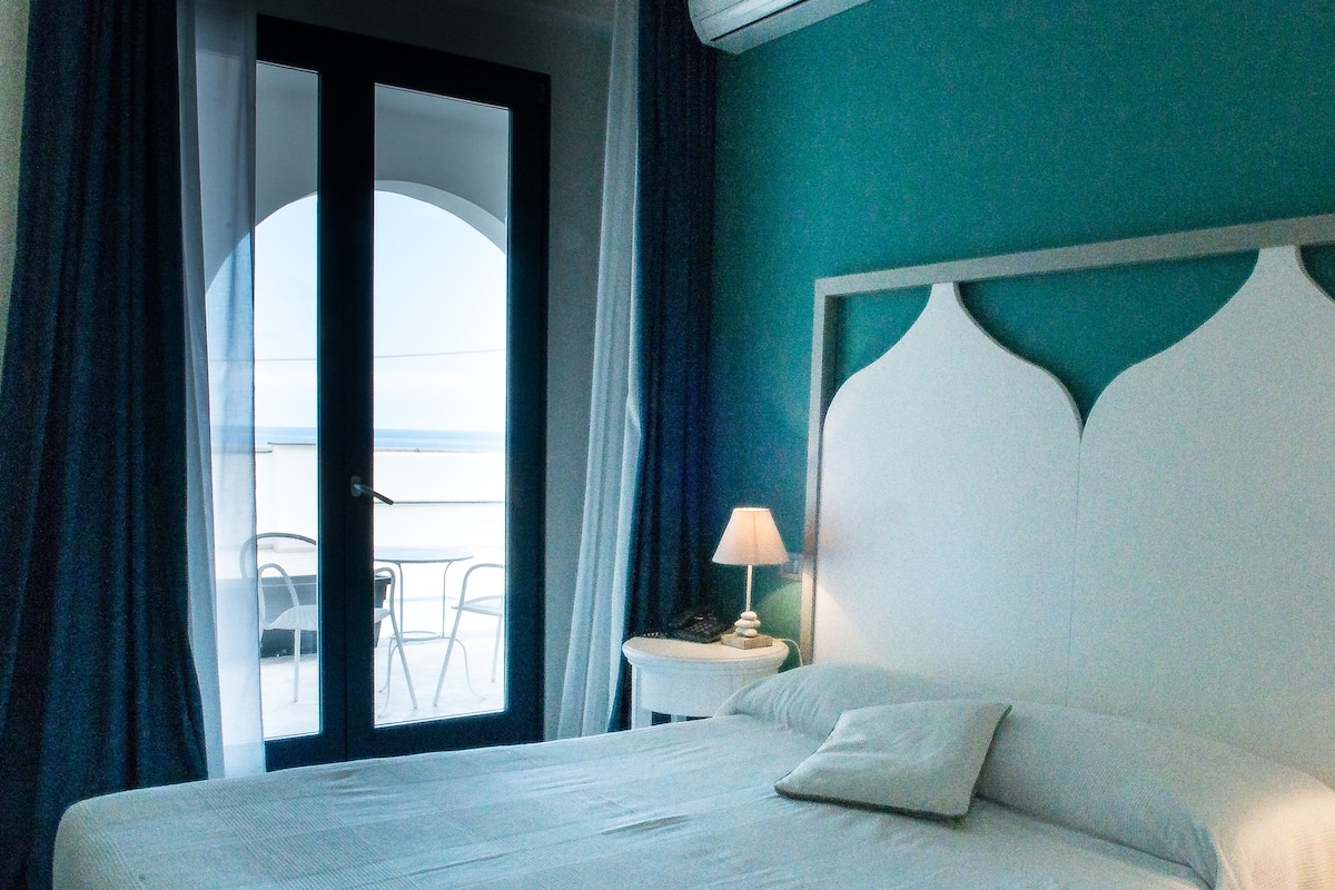 A hotel room with a bed, a nightstand with a lamp, a view of the outdoors through a glass door, and a balcony with chairs.