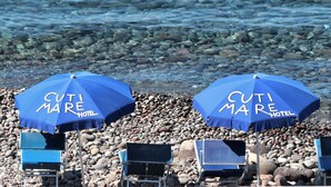 Two blue umbrellas with "CUTI MARE HOTEL" text, a rocky beach, and several blue beach chairs. - Hotel Cutimare (Lipari)