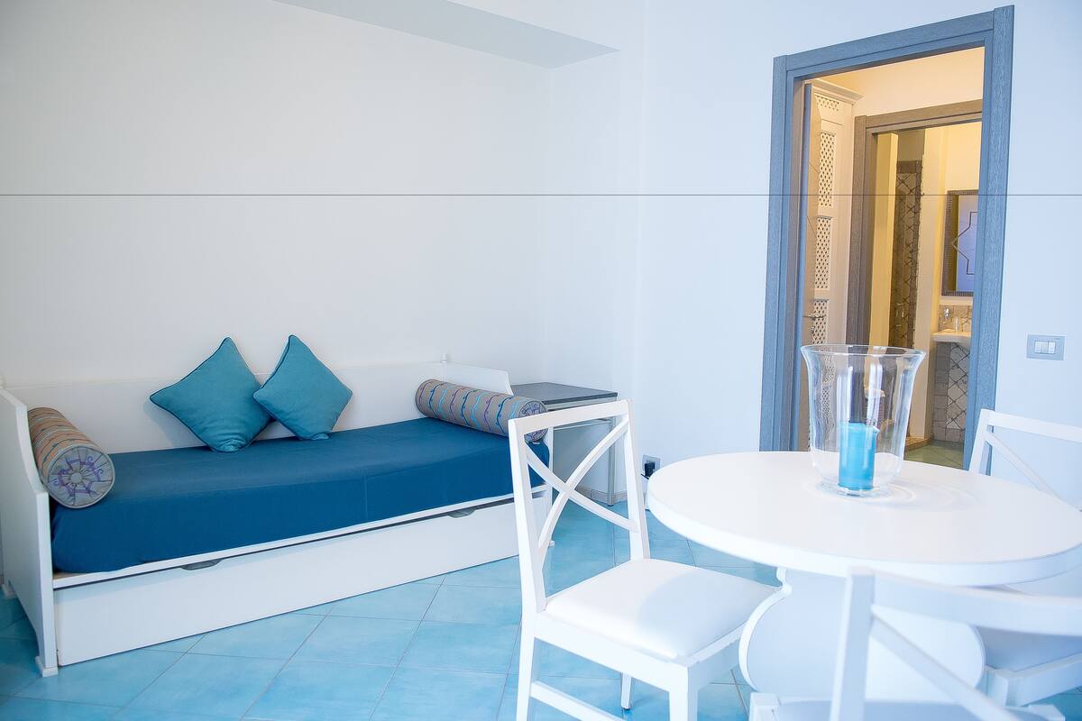 A room with a white sofa with blue cushions, a white dining table with chairs, and a clear glass vase on the table.