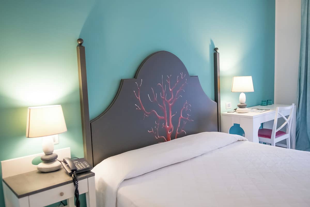 A hotel room with a bed, bedside table with a lamp, a desk with a chair, and a wall with a tree design.
