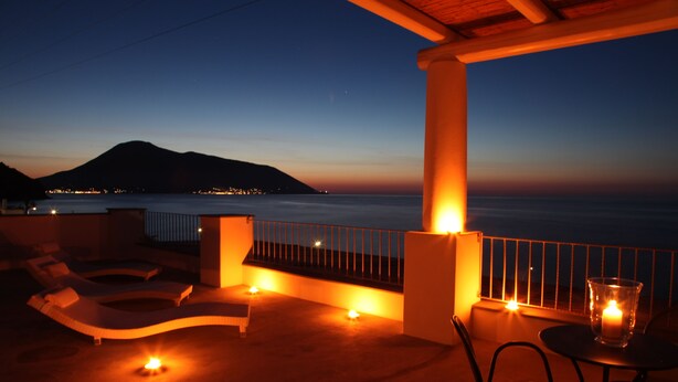A terrace with lounge chairs, a table, and candles, overlooking a calm sea and a mountain at dusk. - Hotel Cutimare (Lipari)