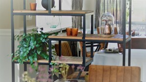 A dining area with a wooden table and chairs, a metal shelf with decorative items, and a potted plant. - Hotel de la Gaichel (Habscht)