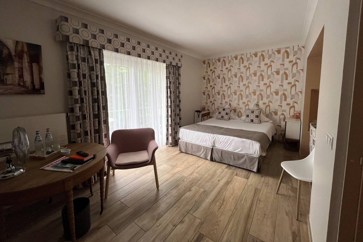 Ein Hotelzimmer mit Holzboden, einem Bett mit gemustertem Kopfteil, einem Schreibtisch mit Stuhl und einem Fenster mit Vorhängen.