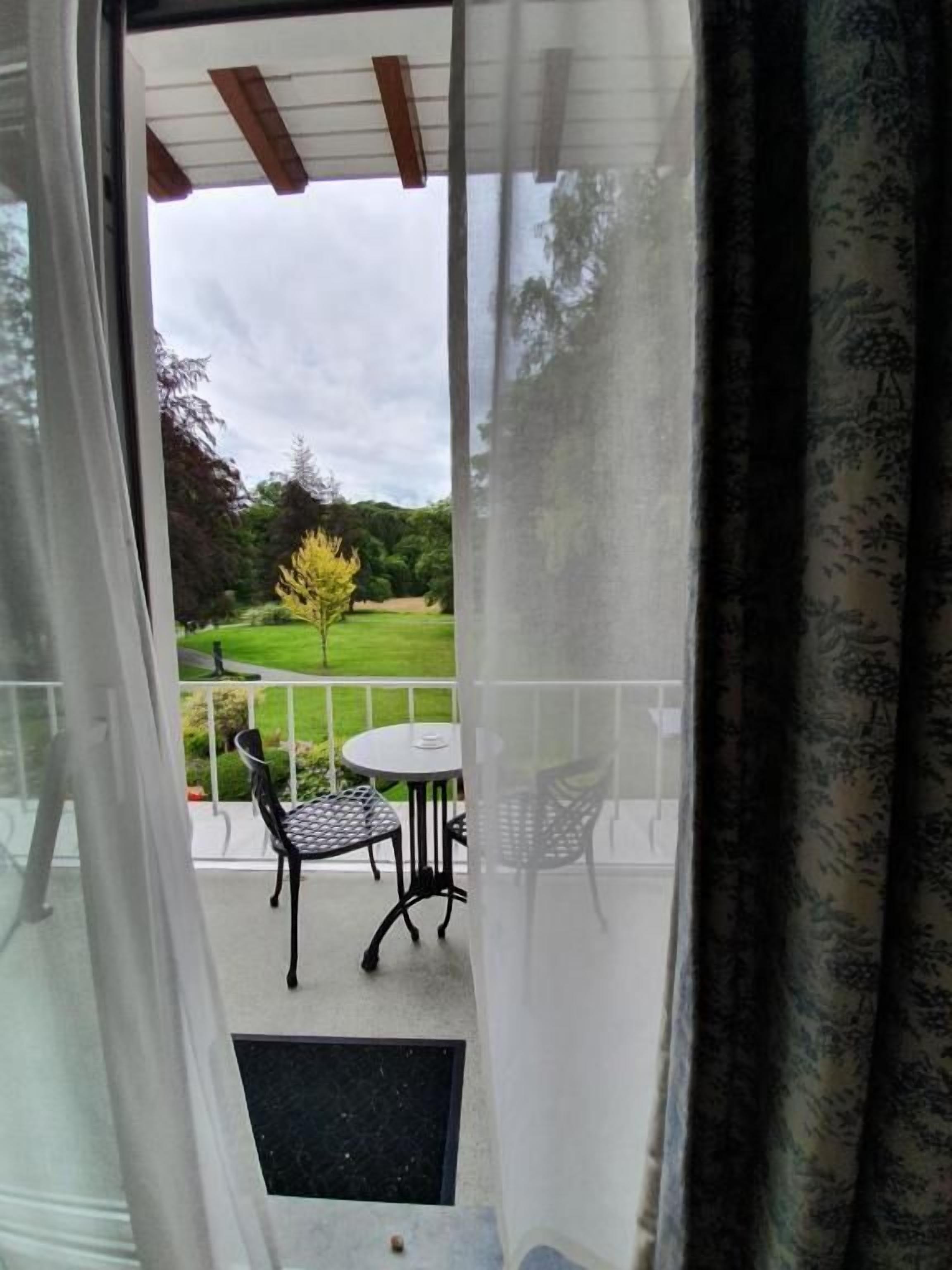 Uitzicht door een glazen deur op een terras buiten met een tafel en stoelen, een gazon en bomen.