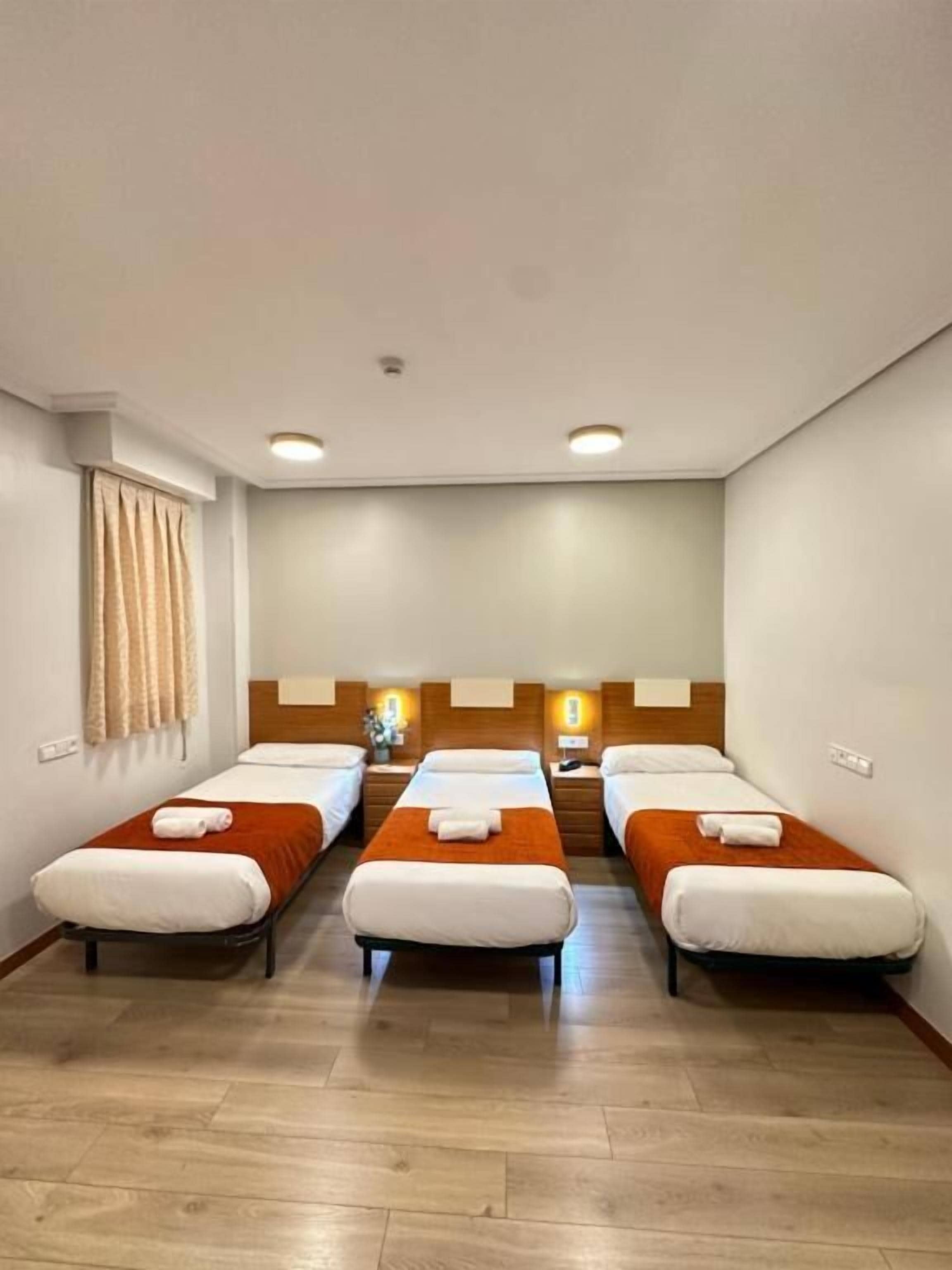 A hotel room with three single beds aligned against the wall, a wooden headboard, and a small table between the beds.