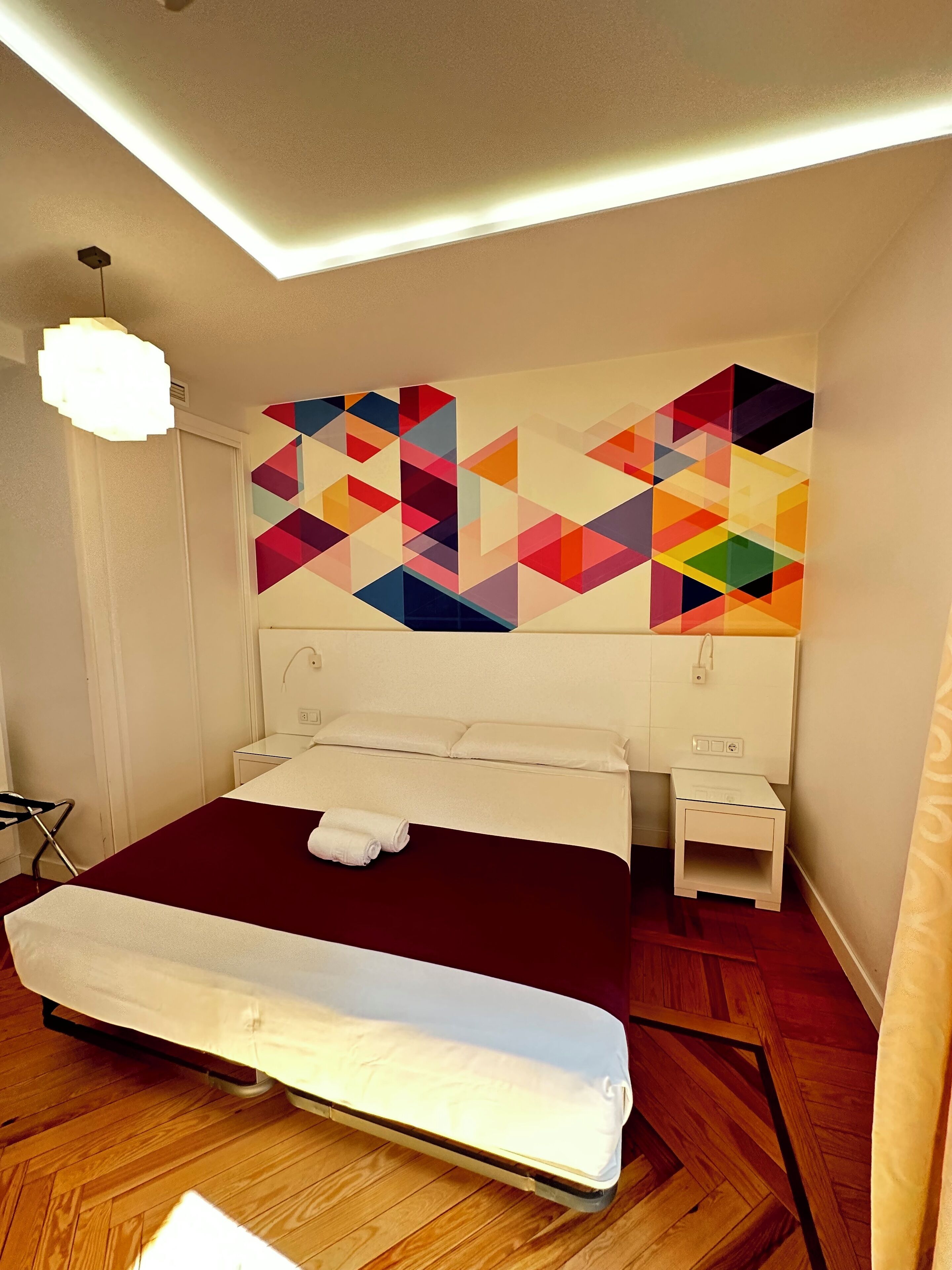 A modern hotel room with a large bed, a bedside table, a nightstand, and a colorful geometric wall art piece.