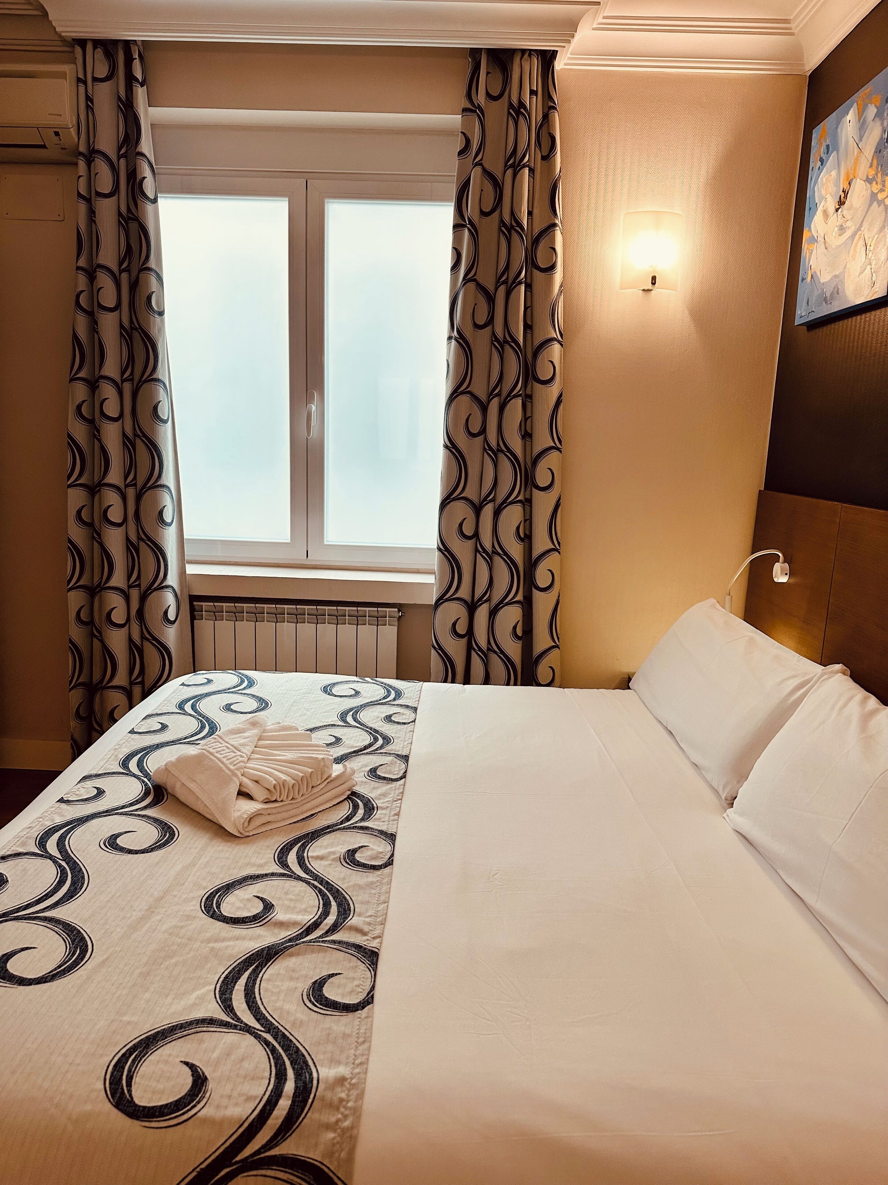 A hotel room with a bed, curtains, a window, a radiator, and a wall-mounted light.
