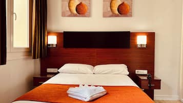 Hotelkamer met houten hoofdeinde, twee bedlampen, een bed met witte lakens, een oranje plaid en twee schilderijen van vazen aan de muur.