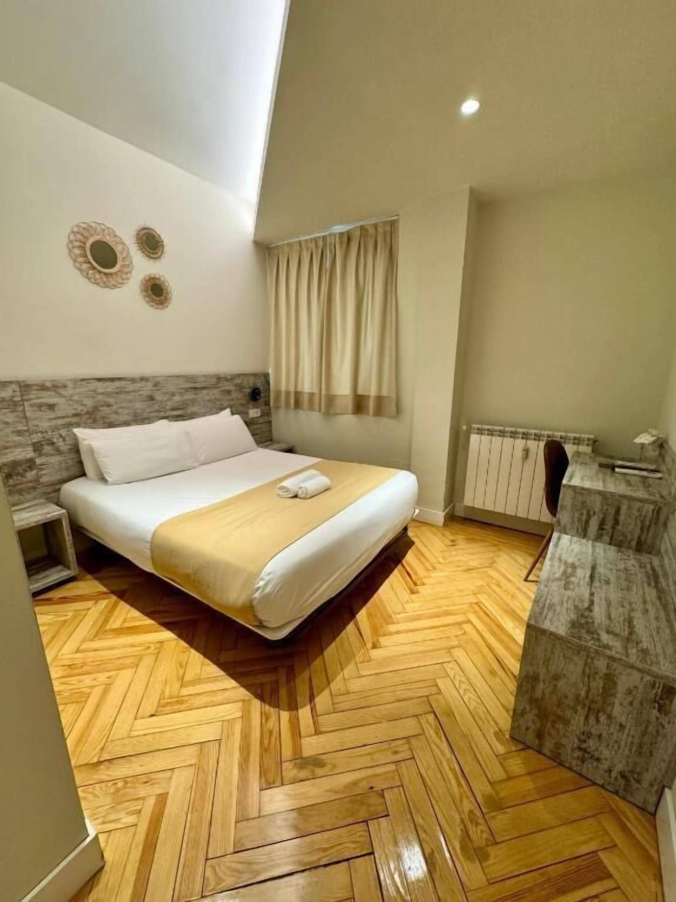 Camera d'albergo con pavimento in parquet a spina di pesce, un letto con lenzuola bianche, una testiera grigia, un comodino e un armadietto grigio.