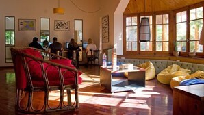 A spacious room with wooden floors, a high ceiling, and large windows allowing natural light. There are several seating areas with cushions and a central table. - El Gualicho Hostel (Puerto Madryn)