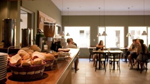 A buffet area with bread baskets and plates on the counter, tables and chairs in the background, and a large window providing natural light. - El Gualicho Hostel (Puerto Madryn)
