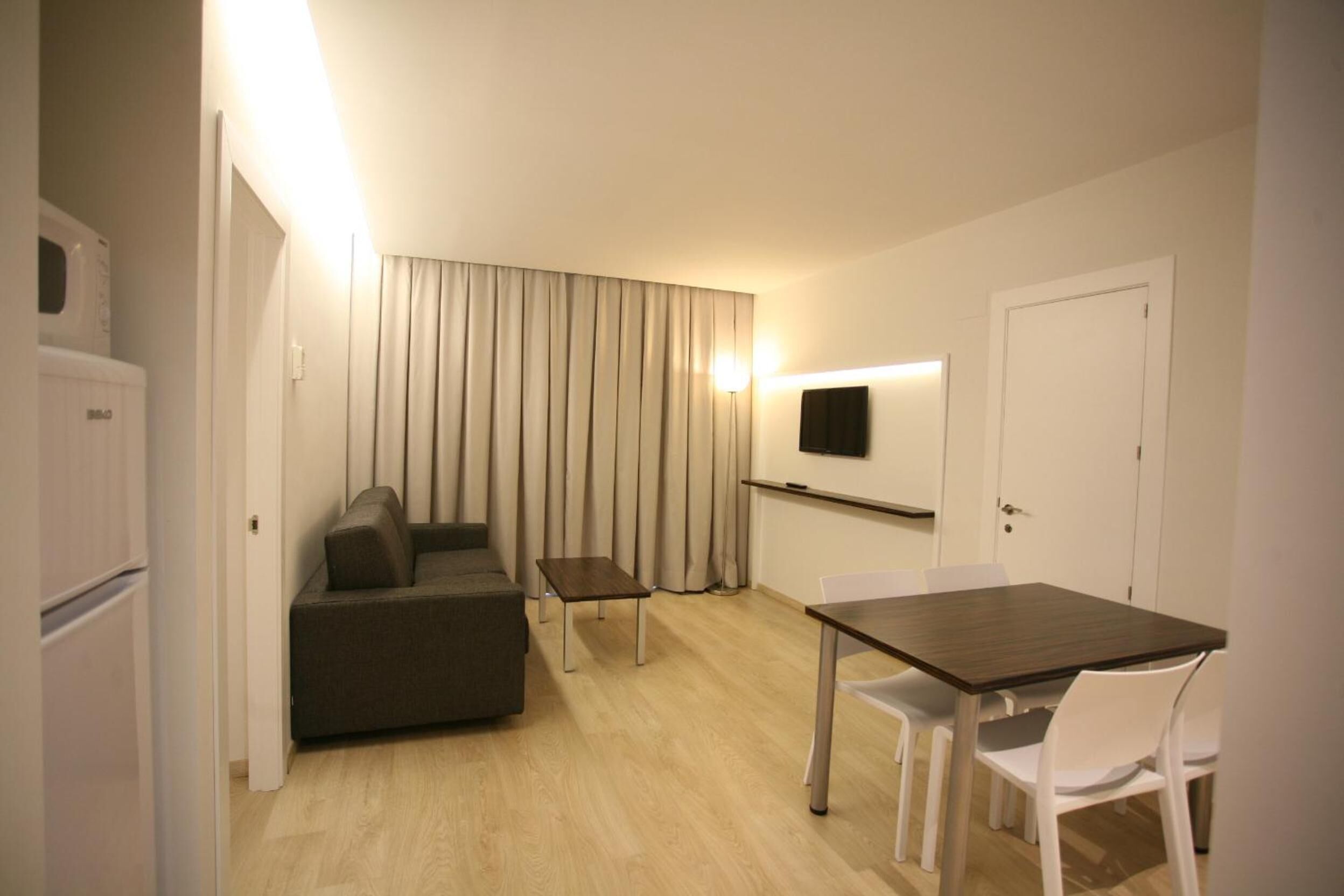 A compact room with a sofa, a small table, a TV mounted on the wall, and a door.