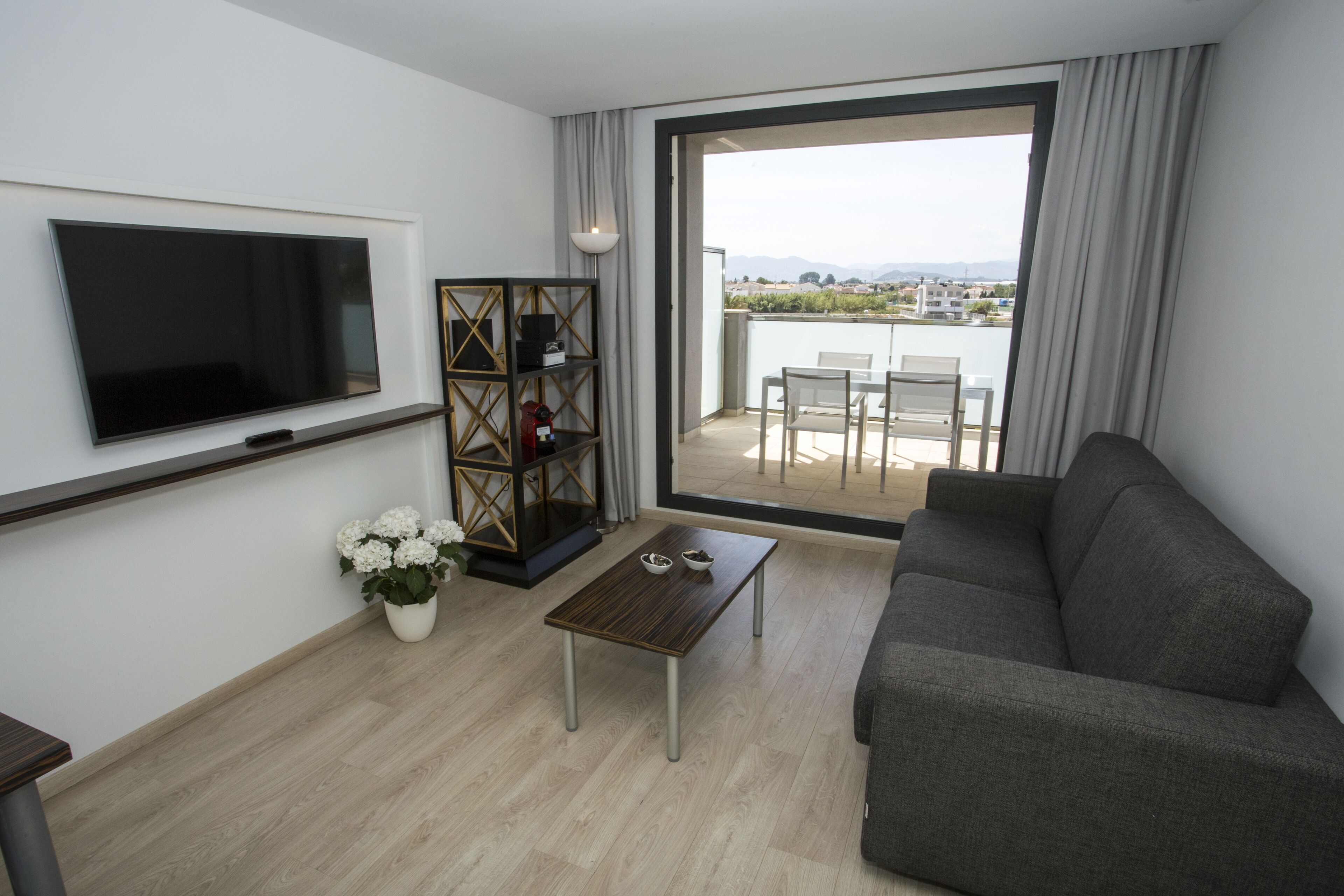 A modern living room with a flat-screen TV, a sofa, a coffee table, and a balcony with a view.