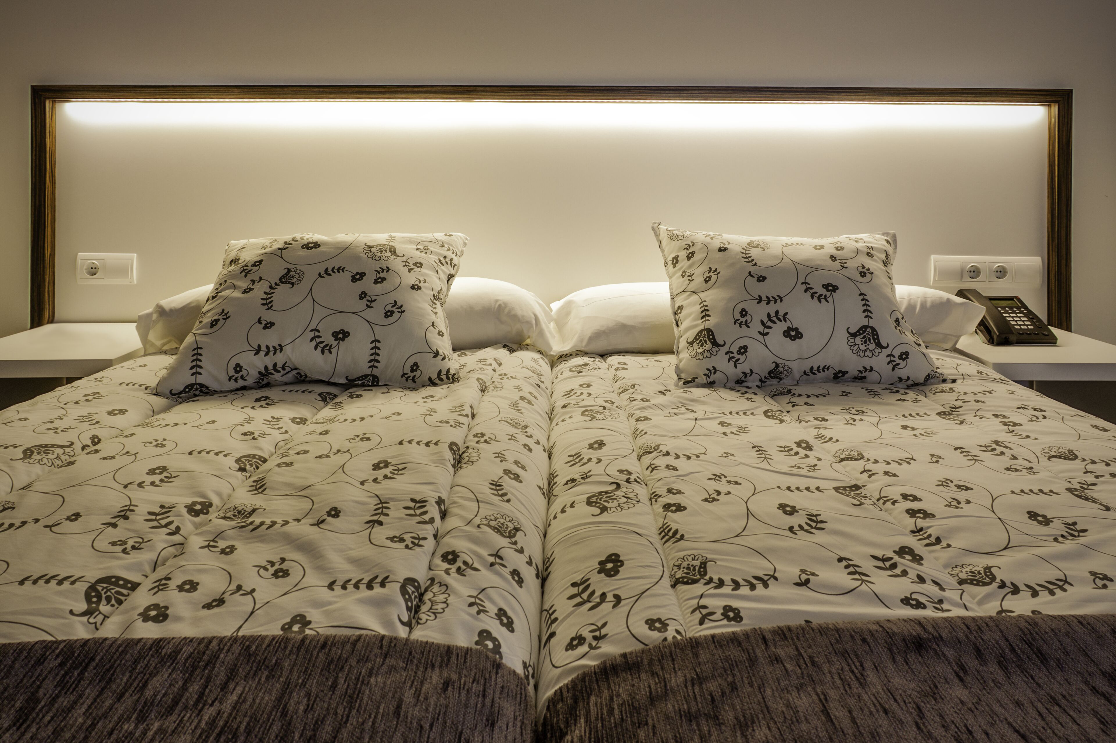 A double bed with floral-patterned bedding, two pillows, a bedside table with a phone, and a wall-mounted light.
