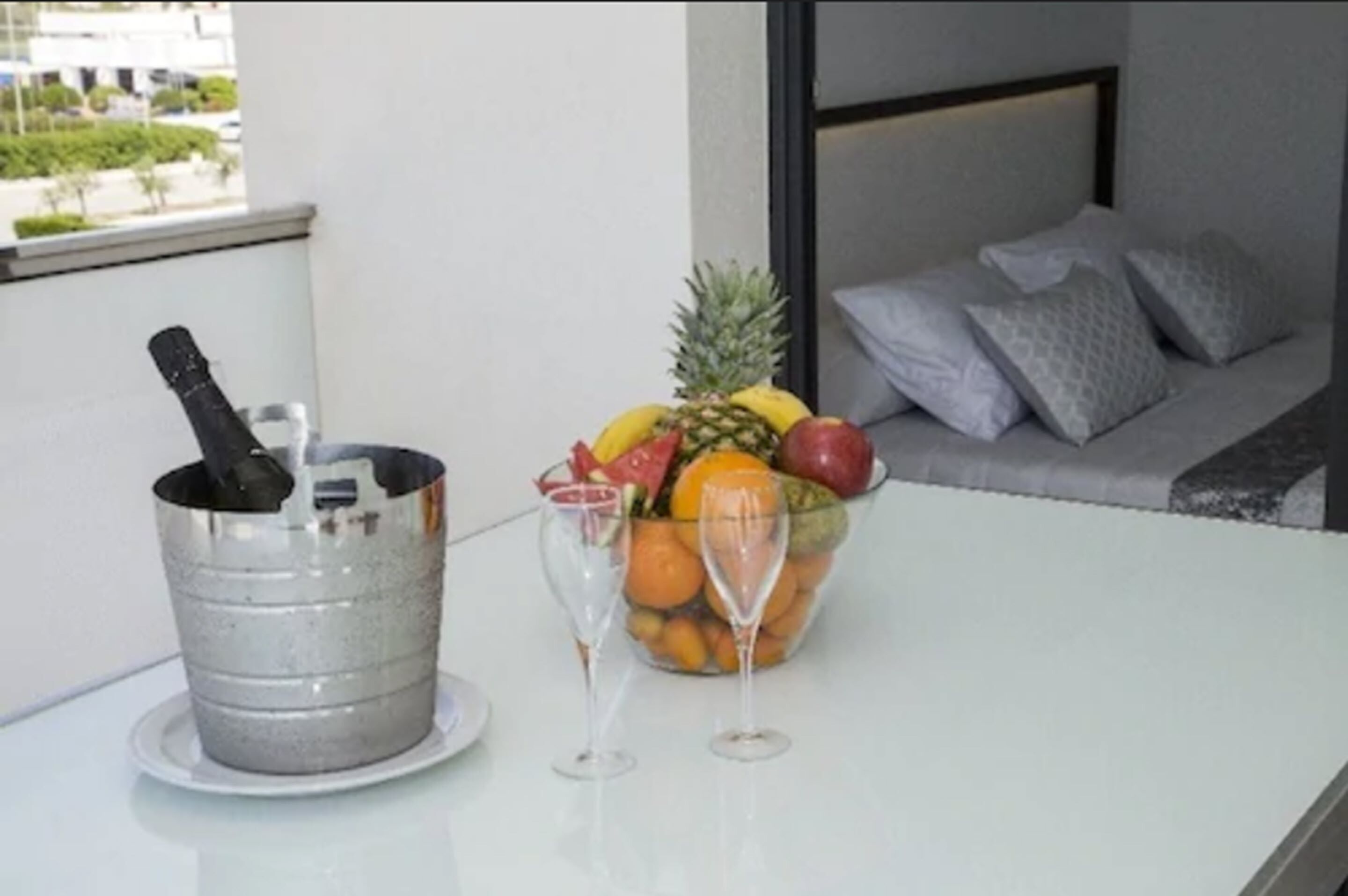 A table with a fruit bowl and two wine glasses, a metal bucket, and a view of a balcony with a sofa and cushions.
