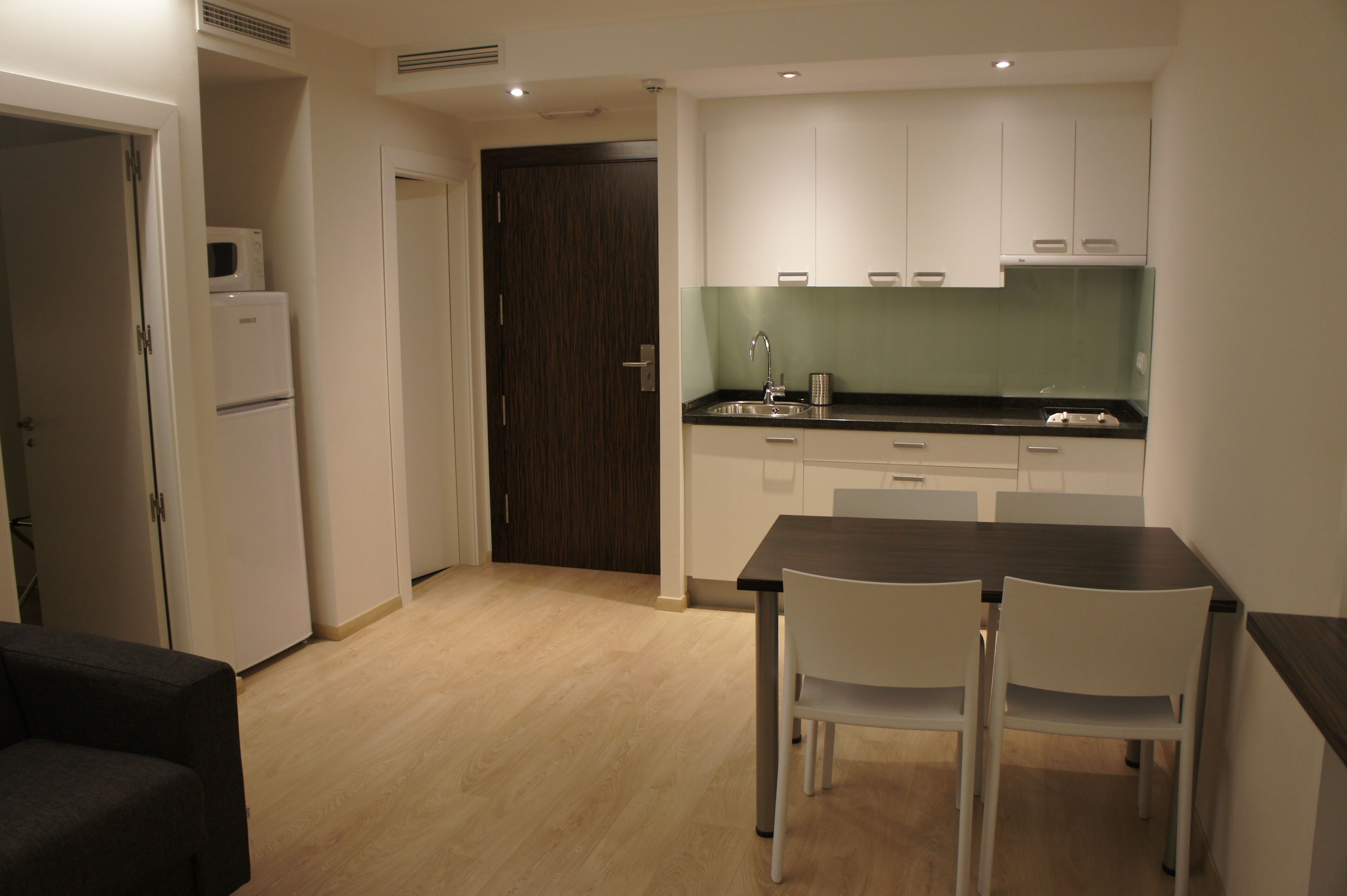 A compact kitchen with white cabinets, a black countertop, and a dining area with two white chairs.