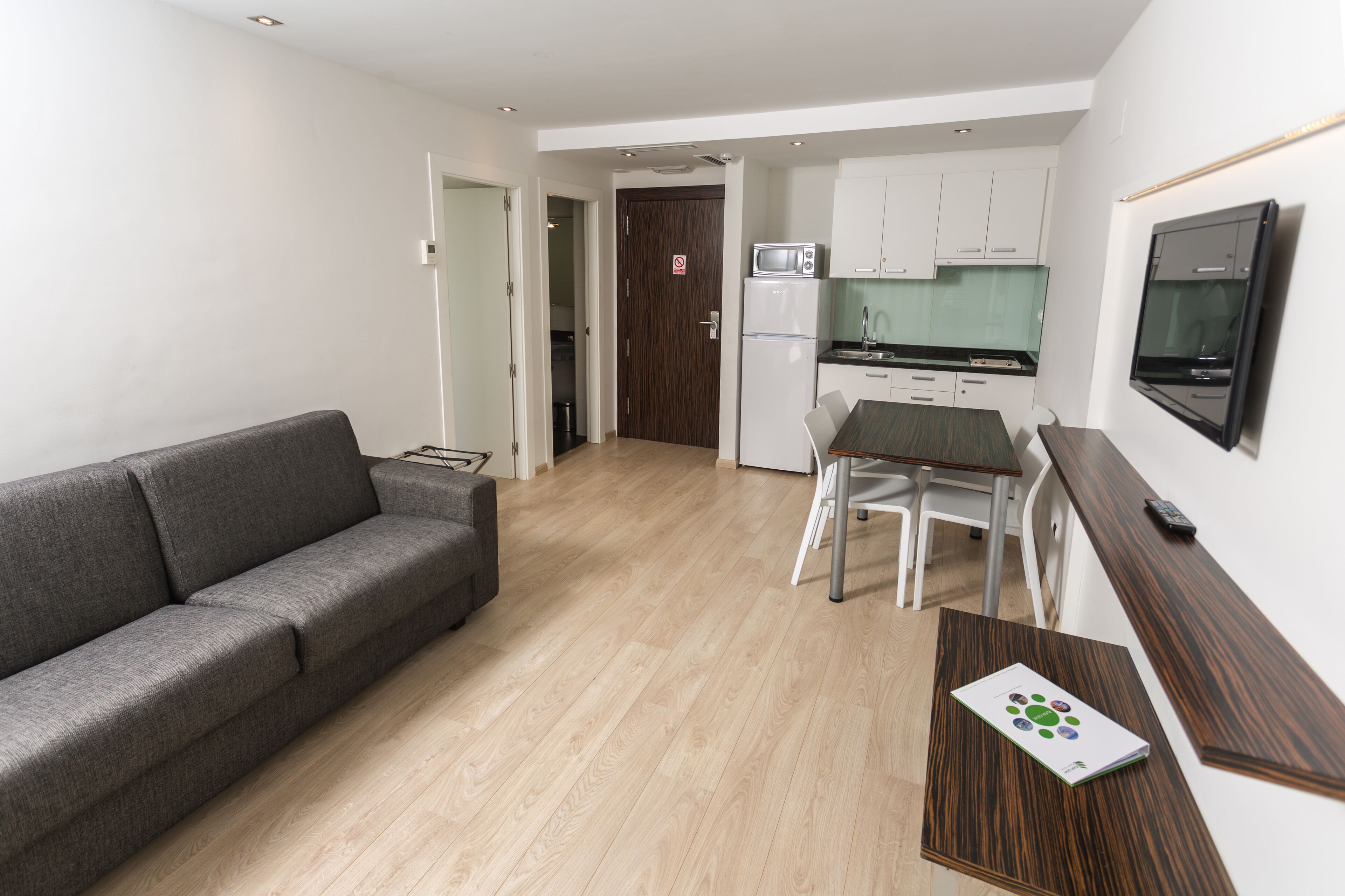 Een modern appartement met een grijze bank, een houten eettafel, een aan de muur bevestigde televisie en een keuken met witte kasten en een magnetron.