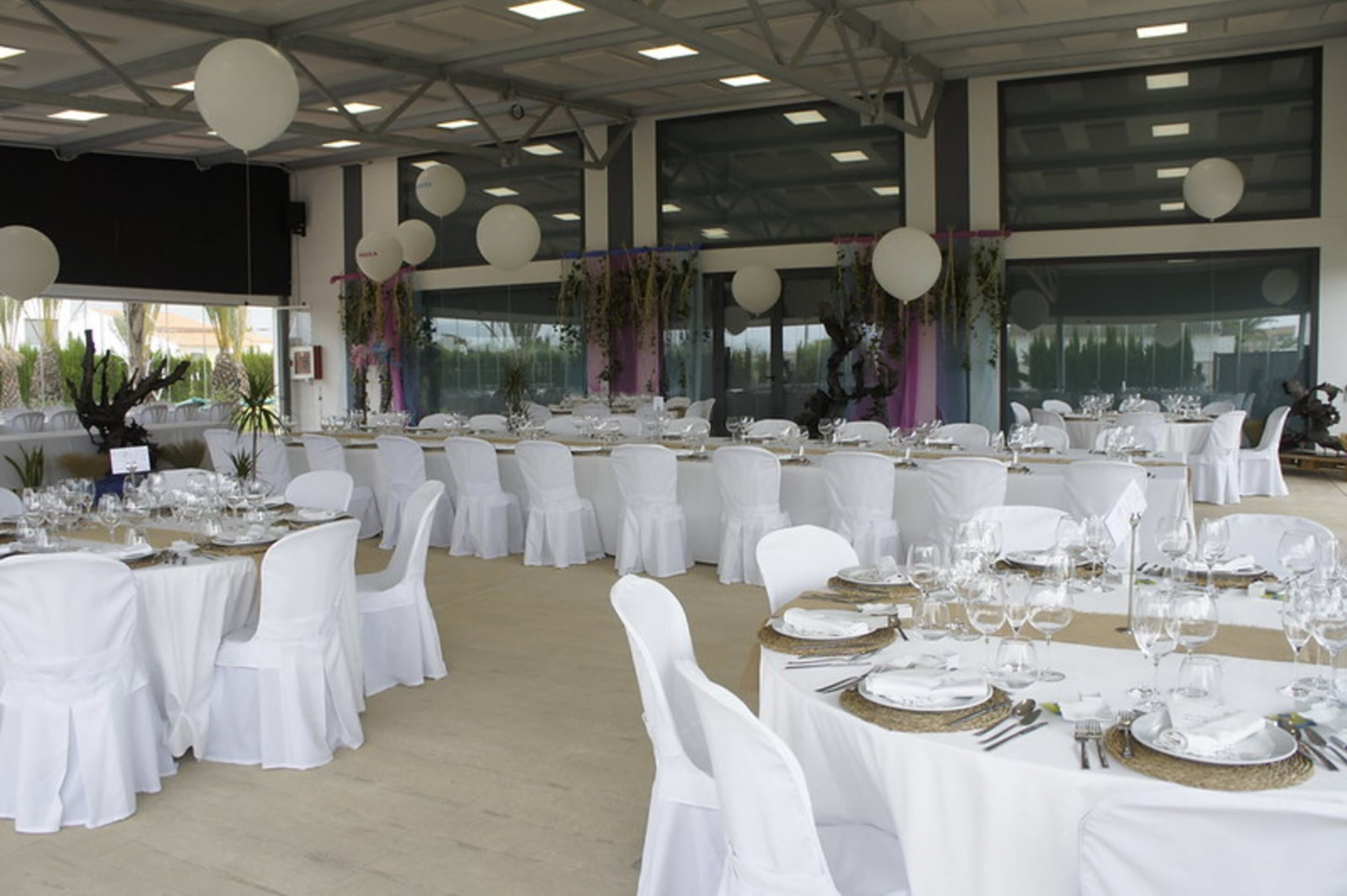 Un salón de banquetes con mesas redondas dispuestas para un evento formal, manteles blancos y sillas con fundas blancas.