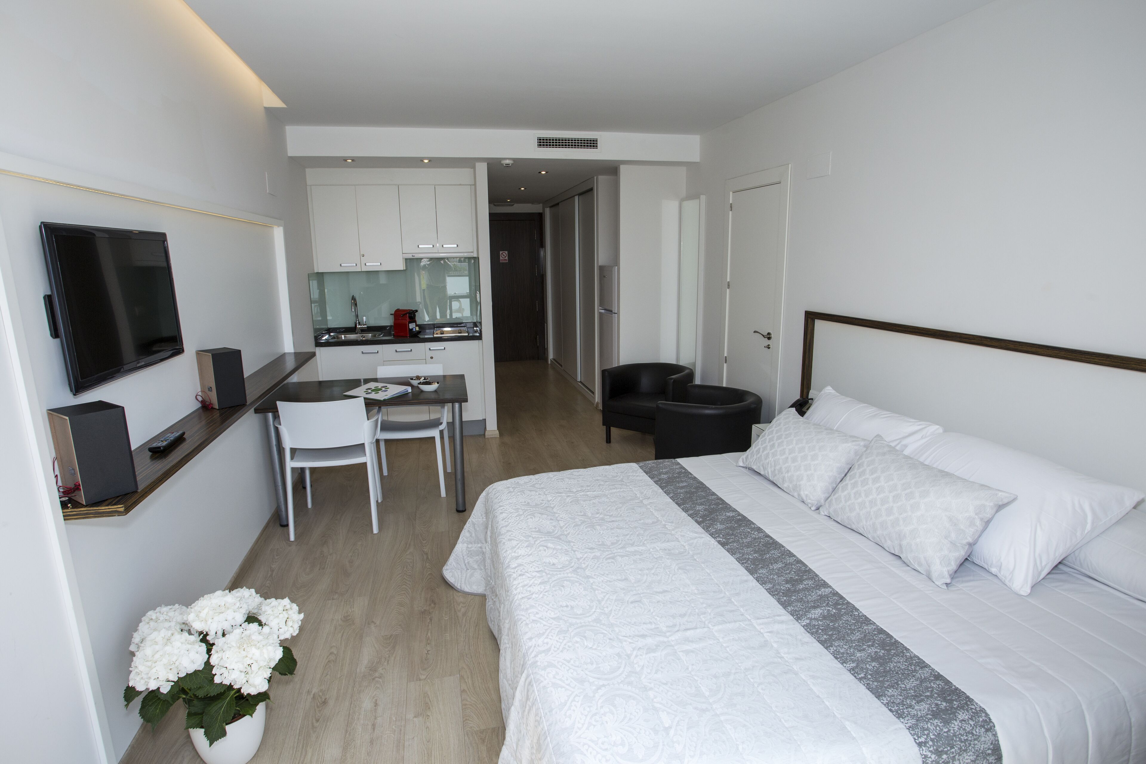 A modern hotel room with a large bed, a desk with a chair, a television, and a kitchenette in the background.
