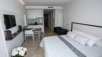 A modern hotel room with a large bed, a desk with a chair, a television, and a kitchenette in the background.