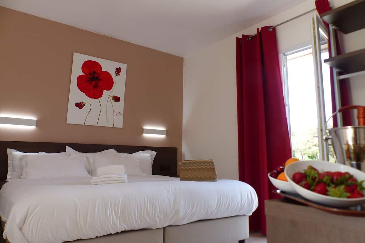 A hotel room with a large bed, a painting of red flowers, a bowl of fruit, and a window with red curtains.