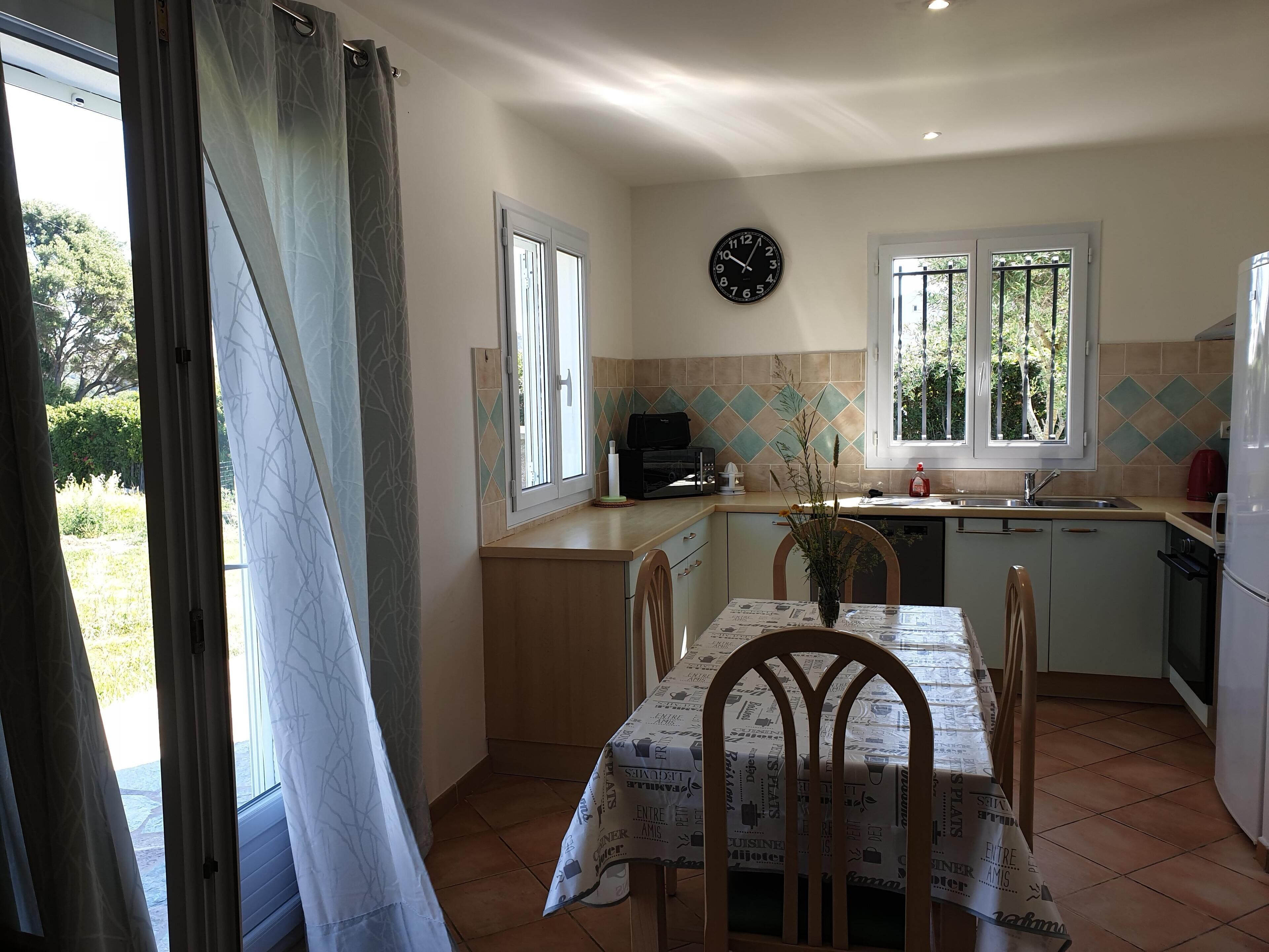 Une cuisine avec une table à manger, des chaises, une horloge et une fenêtre donnant sur un paysage verdoyant.