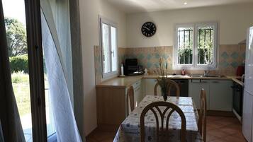 Une cuisine avec une table à manger, des chaises, une horloge et une fenêtre donnant sur un paysage verdoyant.
