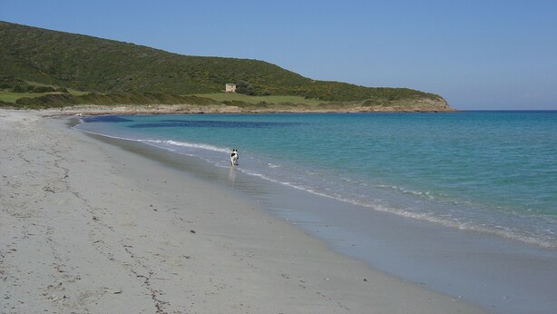 A beach with clear blue water, a sandy shore, and a hill with a building in the background. - Stella Marina (Rogliano)