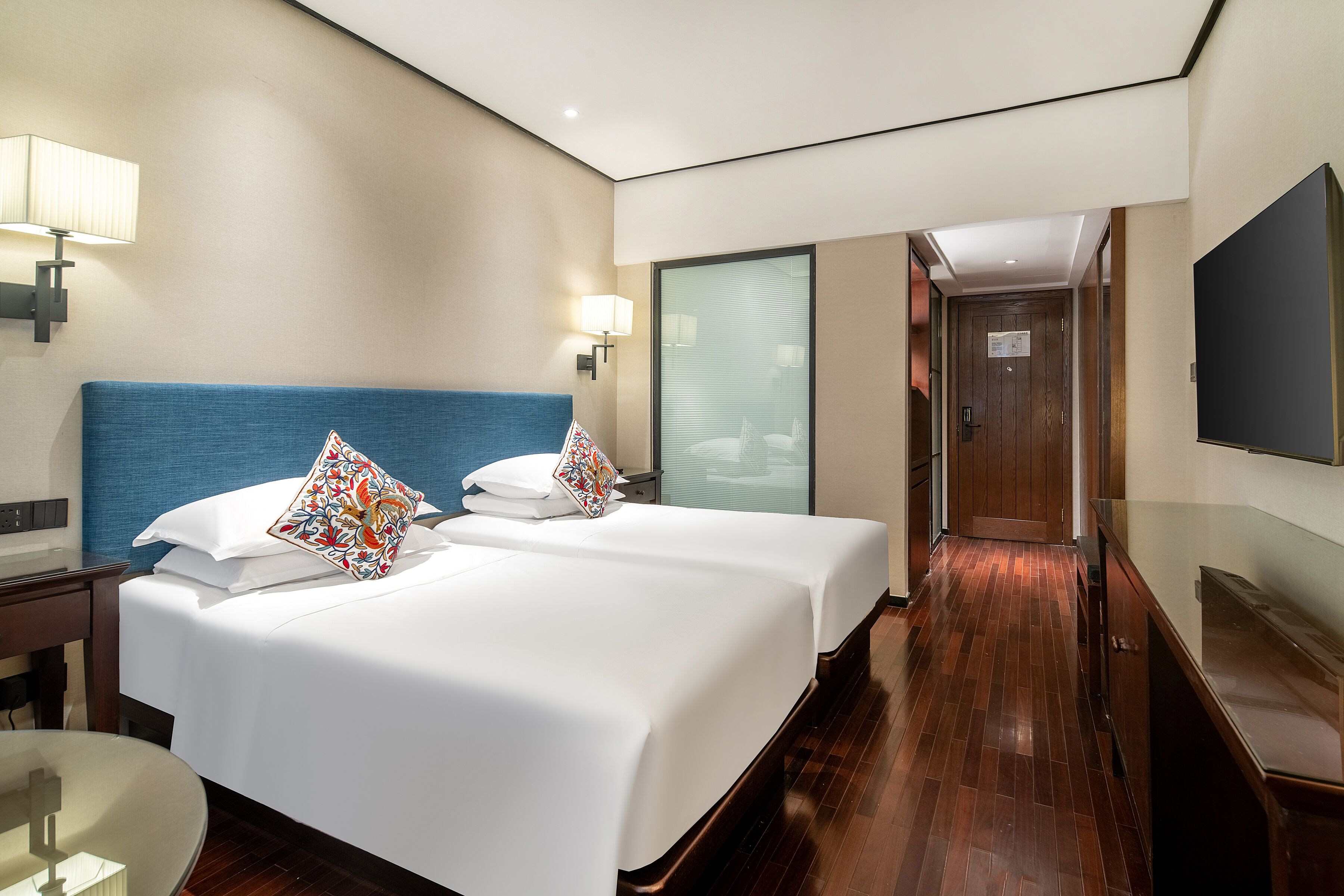 A hotel room with two beds, a desk, a chair, a TV, and a bathroom visible through a glass door.