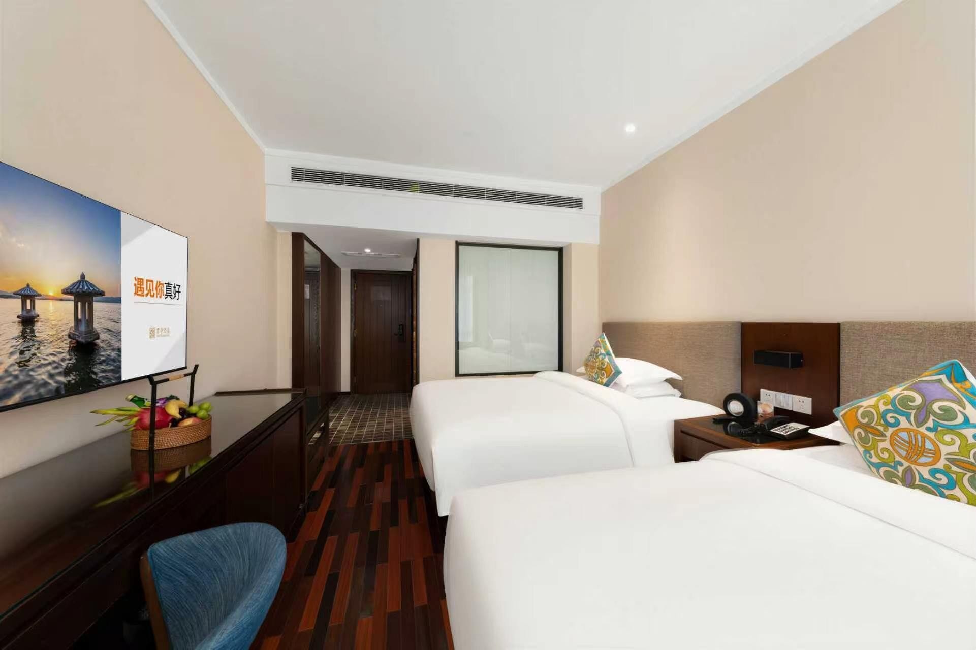 A hotel room with two beds, a desk, a chair, a TV, and a wall-mounted air conditioning unit.