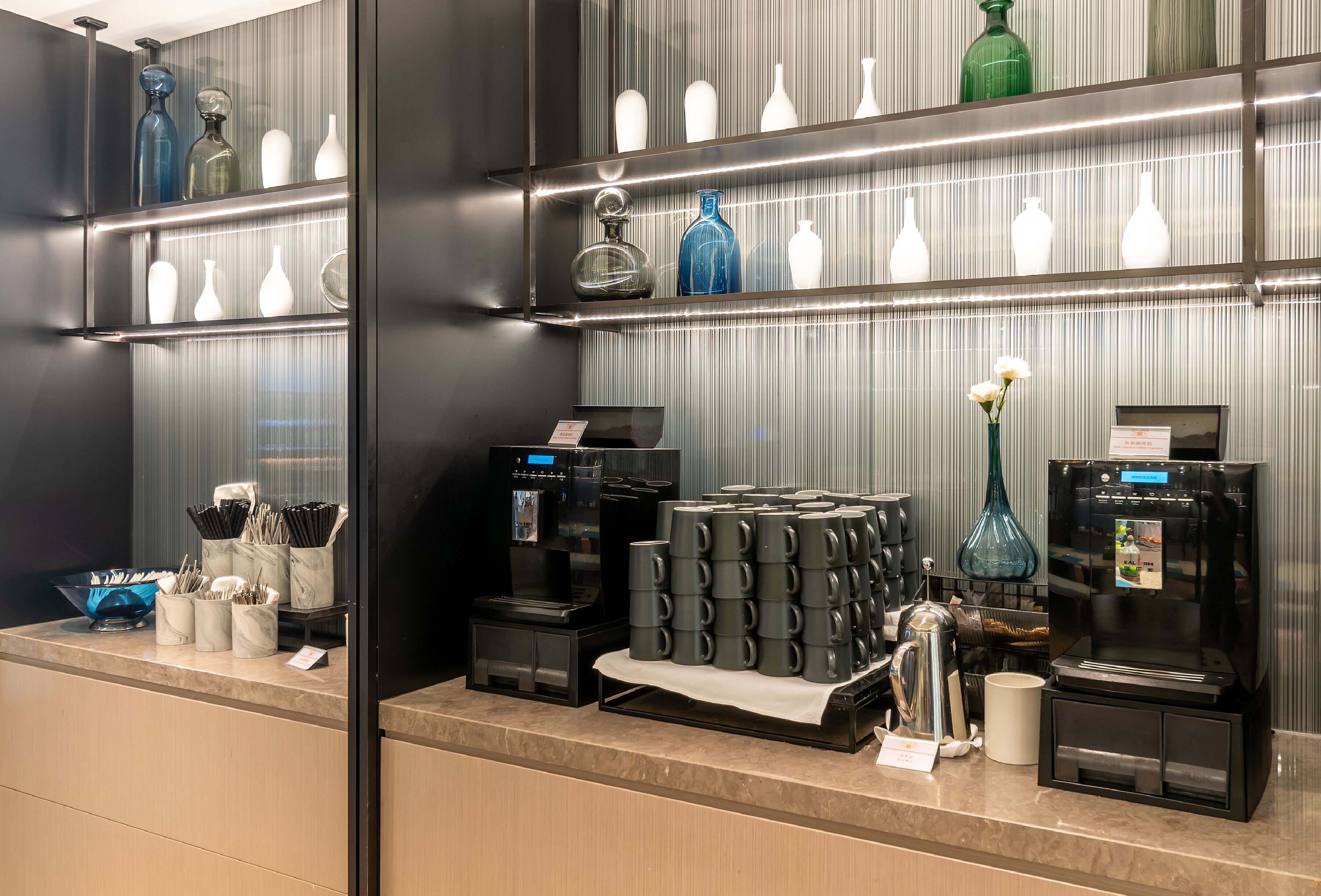 A hotel reception area with a coffee station, shelves with various glassware, and a modern design.